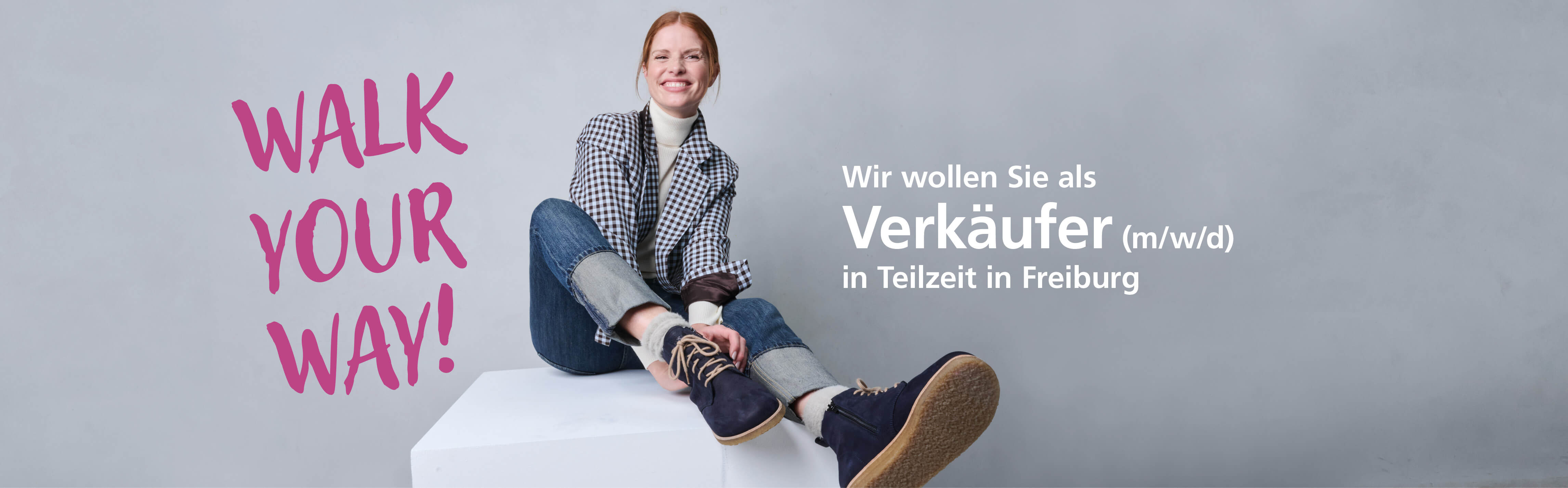Junge Frau sitzt lächelnd in Freizeitkleidung neben Schriftzug 'Walk Your Way'. Gesucht wird ein Verkäufer (m/w/d) in Teilzeit in Freiburg.