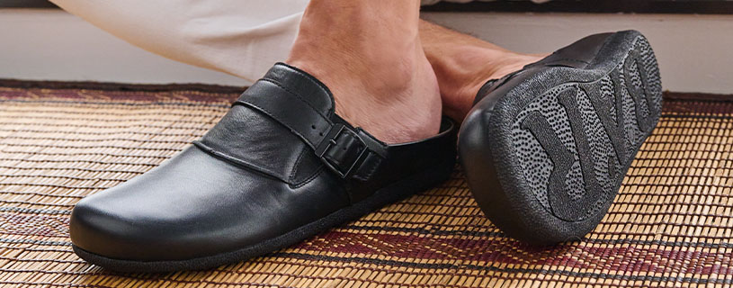 Ein schwarzer Herrenslipper aus Leder mit ergonomischer Sohle auf einer geflochtenen Matte