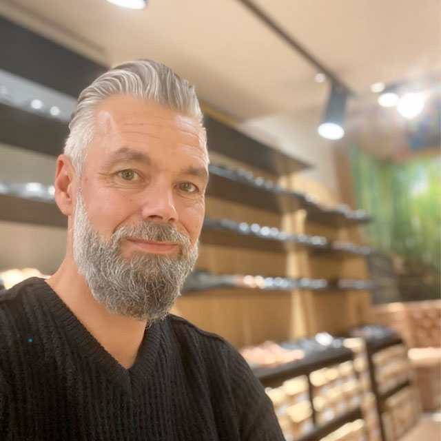 Ein Mann mit grauem Haar und Bart steht vor einem Regal in einem Geschäft und lächelt in die Kamera.